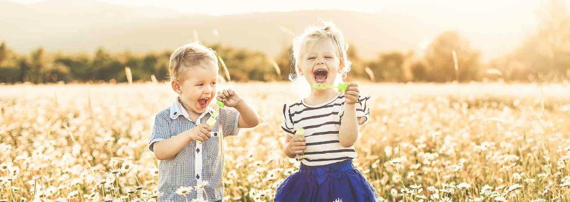 Zwei Kinder spielen auf einer Blumenwiese lachend mit Seifenblasen